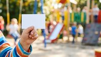 Child's hand holding blank white card at playground with blurred background of play equipment and other children, suitable for advertising, messaging, or promotional campaigns.