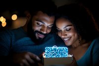 Young African American couple smiling while looking at glowing gift box in dark room, romantic moment with bokeh lights creating magical atmosphere.