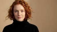 Young redhead woman with curly hair wearing black turtleneck sweater against neutral beige background for fashion and lifestyle content.