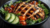 Grilled chicken breast with grill marks served on fresh green salad with cherry tomatoes and cucumber slices in dark bowl, close up view on wooden table.