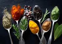 Colorful aromatic spices and herbs in vintage silver spoons on dark slate background, overhead view of turmeric, paprika, pepper, rosemary and bay leaf arrangement.