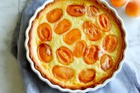 Fresh apricot custard tart with golden fruit halves arranged on creamy vanilla filling in white ceramic baking dish, photographed from above on marble surface.