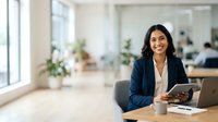 Young South Asian woman in professional business attire working at desk with tablet and laptop in modern office environment with natural light and plants.