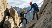 Two climbers with safety gear ascending rocky mountain cliff face with dramatic alpine peaks in background during outdoor adventure expedition.