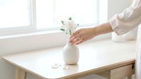 Woman in white shirt arranging white flowers in ceramic vase on wooden table by bright window, minimalist home decor and floral arrangement styling.