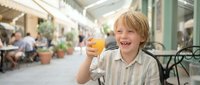 Happy young Caucasian boy enjoying fresh orange juice at outdoor cafe terrace, smiling cheerfully while holding glass, summer vacation lifestyle concept.