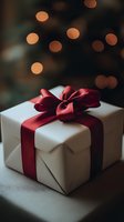 White gift box wrapped with elegant red satin ribbon bow against blurred warm bokeh lights background, creating festive holiday atmosphere.