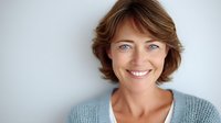 Confident middle-aged woman with bright smile and blue eyes wearing light sweater against white background, perfect for wellness or healthcare marketing.