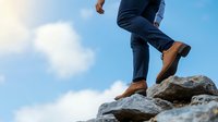 Young Caucasian businessman in navy suit climbing rocky mountain peak against blue sky with clouds, representing leadership and career achievement goals.
