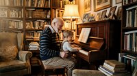 Grandfather and grandson playing piano together in a warm, book-filled study room with vintage furnishings and soft lamp lighting. Intergenerational bonding moment.