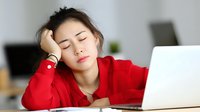Young Asian woman in red shirt showing signs of fatigue or burnout while working on laptop, illustrating workplace stress and mental exhaustion.