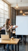 Professional businesswoman presenting at whiteboard in modern office with geometric pendant lights and large windows for corporate training sessions.