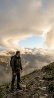Male hiker in dark jacket standing on mountain ridge overlooking misty valley with dramatic cloud formations and alpine peaks at sunrise or sunset.