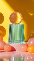 Crystal glass pedestal with transparent sphere balanced on top, surrounded by pink and orange orbs against yellow background. Minimalist luxury setup.
