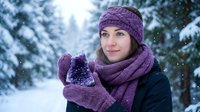 Young Caucasian woman in purple winter knit accessories holds amethyst crystal in snowy forest, snowflakes falling around her.