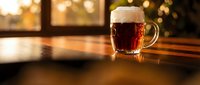 Dark beer mug with foam head on wooden table in warm pub atmosphere with golden bokeh lights creating cozy evening ambiance for hospitality concepts.