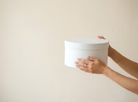 Person with medium skin tone holding white cylindrical gift box against neutral background, suitable for holiday, celebration, and retail marketing materials.