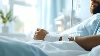 Patient lying in hospital bed with IV drip, close-up view of hands resting on white sheets. Medical care setting shows modern treatment room with natural lighting.