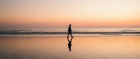 Silhouette of male surfer carrying surfboard walking on wet beach sand at golden sunset, reflected in water, ready for ocean waves and water sports activity.