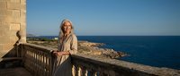 Mature blonde woman in beige coat standing on stone balcony overlooking blue Mediterranean sea and rocky coastline during golden hour sunset.