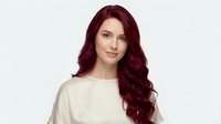 Young Caucasian woman with long wavy burgundy red hair wearing cream sweater smiling at camera against light background for beauty and fashion content.