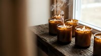 Lit candles in glass jars arranged on wooden shelf by window, releasing aromatic smoke for home fragrance, relaxation, and interior decoration purposes.