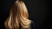 Back view of long blonde layered hair with natural highlights flowing against dark background, demonstrating professional hairstyling and healthy hair movement.