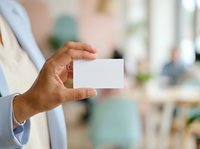 Man in light blue shirt holding blank white business card in office environment with blurred background, suitable for corporate branding and professional networking materials.