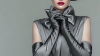 Elegant woman in silver satin dress and matching gloves with dramatic bow detail, luxury fashion portrait on neutral background for editorial use.