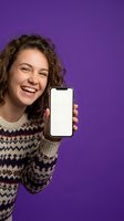 Happy young woman with curly hair showing blank smartphone screen on purple background for app presentation and mobile marketing campaigns.
