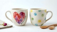 Young woman with South Asian ethnicity, wearing casual home attire, holding two ceramic mugs with watercolor heart and polka dot designs, styled for lifestyle and home decor content.