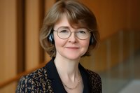 Professional middle-aged Caucasian woman wearing round glasses and lace blouse smiling warmly at camera, business portrait with soft natural lighting and blurred office background.