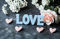 Blue wooden letters spelling LOVE with pink heart decorations and delicate white roses on dark textured background, romantic wedding or valentine composition.