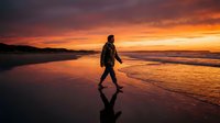 Silhouetted male figure walking along sandy beach during dramatic golden sunset with vibrant orange and purple sky, ocean waves, and coastal landscape for travel and lifestyle content.