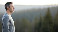 Middle-aged man with gray hair and light skin tone, wearing casual gray shirt, gazing thoughtfully at forested mountain landscape. Suitable for wellness, lifestyle, and contemplation themes.