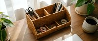 Wicker desk organizer with multiple compartments holding office supplies on wooden table with plants and pottery in bright natural light workspace.