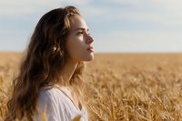 Young woman with long brown hair in white dress standing in golden wheat field during sunset, side profile view showing natural beauty and serenity.