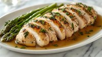 Sliced herb-seasoned chicken breast with butter sauce and fresh asparagus spears served on white oval plate, close up view of gourmet dinner presentation.