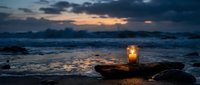 Glowing lantern on driftwood at ocean shore during dramatic sunset with waves and moody sky creating peaceful coastal atmosphere for meditation and relaxation.