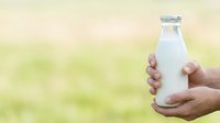 Hand holding glass bottle of fresh milk against blurred green nature background, suitable for dairy product advertising and organic food marketing campaigns.