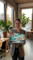 Young woman with brown hair and fair complexion wearing a beige blazer, displaying a tablet with colorful abstract wave graphics in a modern loft office with large windows, potted plants, and city views.