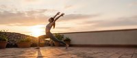 Young athletic woman practicing yoga warrior pose on rooftop terrace during golden hour sunset with arms raised skyward for mindfulness and wellness.