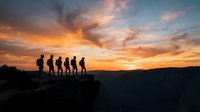 Silhouettes of six hikers standing on a mountain ridge at sunset, gazing at dramatic orange and golden sky with clouds, representing adventure, teamwork, and outdoor exploration.