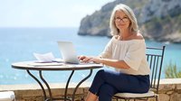 Mature blonde businesswoman working on laptop at seaside cafe terrace with ocean view. Remote work and digital nomad lifestyle concept for professionals.