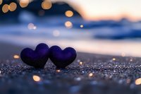 Two purple hearts on sandy beach at sunset with golden bokeh lights creating romantic atmosphere for Valentine's Day and love concepts.