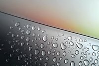 Water drops on dark metallic surface against blurred colorful background, creating natural abstract pattern with light reflection and condensation effect.