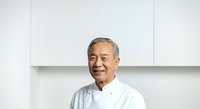 Senior Asian male with gray hair wearing white button-up shirt, smiling at camera in bright office or classroom setting with white wall background.