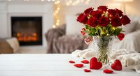 Romantic red roses in crystal vase with scattered heart petals on white table, cozy living room with fireplace and warm lights creating intimate atmosphere.