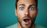 Caucasian man with beard expressing extreme surprise with wide open blue eyes and mouth against turquoise background, showing shock and amazement emotion.