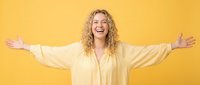 Woman with curly blonde hair, Caucasian ethnicity, wearing pale yellow blouse, arms spread wide in celebratory gesture against yellow background, expressing joy and success.